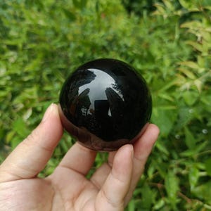 Può includere: Una sfera di ossidiana nera lucidata, che riflette l'ambiente circostante. La superficie liscia e lucida della sfera contrasta con il fogliame verde sullo sfondo. Un oggetto decorativo unico.