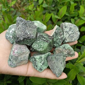 May include: A handful of rough, uncut ruby in zoisite stones. The stones are a mix of dark green and black, with flecks of pink and red. The natural gemstones are held in a hand, with green foliage in the background.