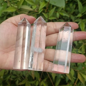 May include: Three clear quartz crystal points, also known as wands, are held in a hand. The points are smooth and polished, with a slight shimmer.