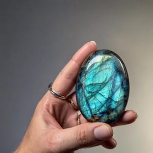 May include: An oval-shaped labradorite gemstone featuring a polished, smooth surface with iridescent flashes of vibrant electric blue and deep teal, contrasted by dark, natural mineral inclusions and fine internal fractures.