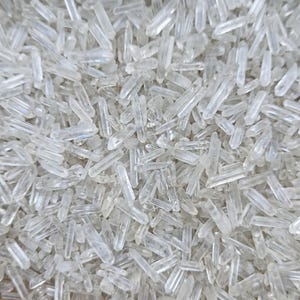 Op de afbeelding: Een close-up van een verzameling heldere kwartskristalpunten. De kristallen zijn van verschillende grootte en vorm, met scherpe, puntige uiteinden. Ze zijn dicht op elkaar geplaatst, waardoor een textuur ontstaat.