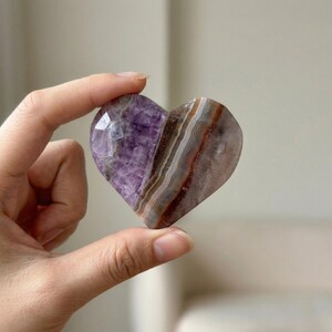 May include: A heart-shaped crystal with a polished surface. The crystal features a blend of purple, brown, and white hues, with visible layers and patterns. The heart is held between fingers, showcasing its shape and color variations.