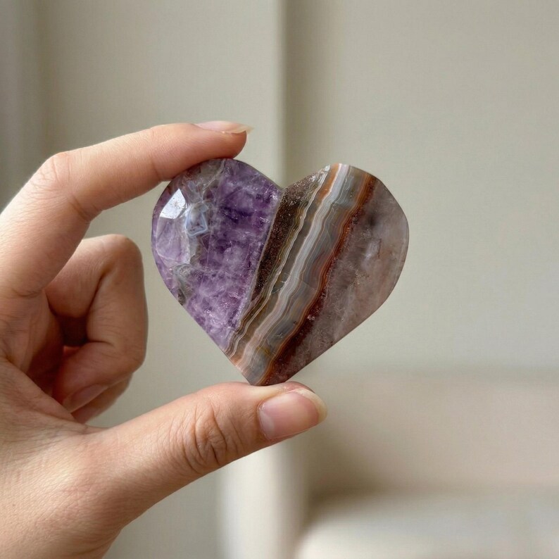 May include: A heart-shaped crystal with a polished surface. The crystal features a blend of purple, brown, and white hues, with visible layers and patterns. The heart is held between fingers, showcasing its shape and color variations.