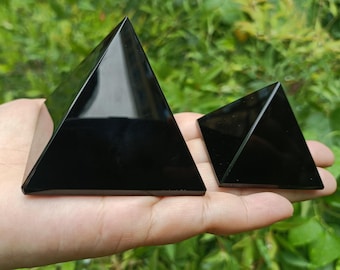 Könnte beinhalten: Zwei schwarze Obsidianpyramiden in unterschiedlichen Größen, eine größer als die andere, werden in einer Hand vor einem grünen, belaubten Hintergrund gehalten.