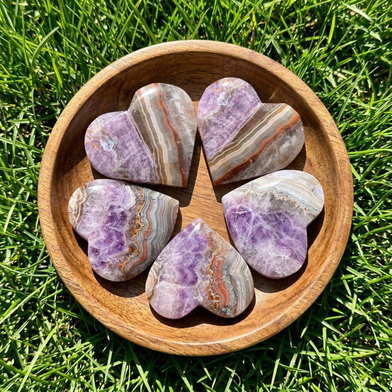 May include: Five heart-shaped amethyst stones with brown and white banding are arranged in a wooden bowl. The bowl sits on a bed of green grass, creating a natural and earthy aesthetic. The stones have a polished finish.