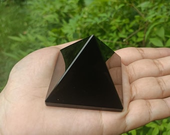 Könnte beinhalten: Eine polierte schwarze Obsidianpyramide mit flacher Basis. Die Pyramide wird in der Hand einer Person gehalten, mit einem grünen, blättrigen Hintergrund. Die Seiten der Pyramide sind glatt und reflektieren das Licht.
