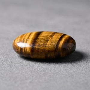 以下が含まれることがあります： 磨かれた楕円形のタイガーアイの宝石。石は、濃い茶色の帯を持つ豊かな金茶色を示し、縞模様を作り出しています。表面は滑らかで反射性があり、わずかな光沢があります。