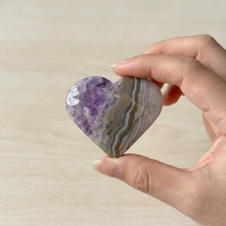 May include: A heart-shaped stone with a polished surface. The stone features bands of purple, gray, and white. The heart is held in a hand, showcasing its natural colors and textures. A unique decorative item.