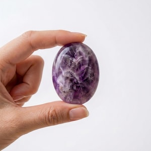 Puede incluir: Una piedra de amatista ovalada y pulida, sostenida entre los dedos sobre un fondo blanco. La piedra muestra una mezcla de tonos morados oscuros y blancos, con una superficie brillante. Esta gema natural se utiliza probablemente con fines decorativos o espirituales.