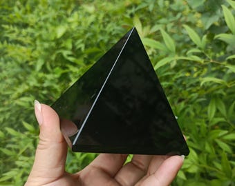 Könnte beinhalten: Ein schwarzer Obsidian-Pyramidenkristall, eine Art Edelstein, wird in einer Hand gehalten. Die Pyramide ist glatt und poliert.