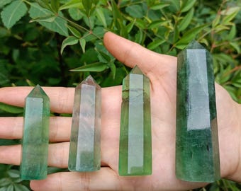 May include: Four green fluorite crystal points of varying sizes are displayed in a hand. The crystals range in color from clear to deep green, with visible internal structures. The crystals are polished and have pointed tops.