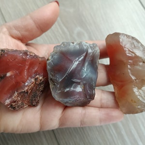 May include: Three agate stones in various shades of red, brown, and white. The stones are rough and have a natural, unpolished look.