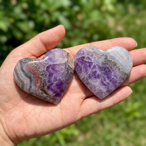 May include: Two heart-shaped amethyst stones with purple, gray, and white patterns. The stones are held in a hand, showcasing their polished surfaces and natural color variations. The heart-shaped stones are a unique gift.