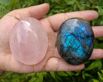 May include: Two polished gemstones: a smooth, oval rose quartz and a labradorite. The rose quartz is a soft pink, while the labradorite displays blue and teal iridescence. The stones are held in a hand against a backdrop of green foliage.