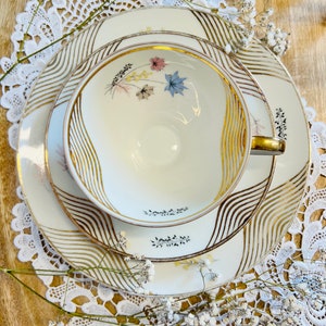 Könnte beinhalten: Ein weißes Tee-Set mit goldenen Wellenmustern und floralem Design. Das Set befindet sich auf einer weißen Spitzenunterlage.