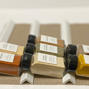May include: A set of eight glass spice jars with black lids and white labels. The labels list the spices: Paprika, Garlic Powder, Onion Powder, Italian Seasoning, Cinnamon, Chili Powder, and Curry. All labels state "All Natural".