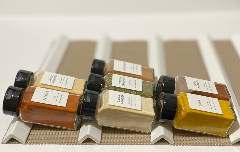 May include: A set of eight glass spice jars with black lids and white labels. The labels list the spices: Paprika, Garlic Powder, Onion Powder, Italian Seasoning, Cinnamon, Chili Powder, and Curry. All labels state "All Natural".