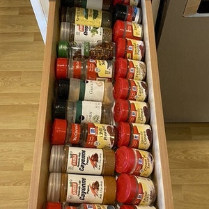 May include: A drawer filled with various spices in small glass jars with red lids. The jars are labeled with the names of the spices, such as "Oregano", "Cayenne", "Garlic", "Fennel", "Paprika", "Nutmeg", "Cinnamon", "Cloves", "Allspice", "Bay Leaf", "Chili Powder", "Cream of Tartar", and "Ground Cloves".