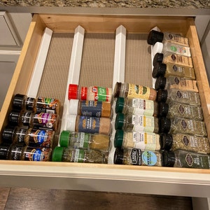 May include: A drawer organizer with white dividers holds a variety of spices in glass jars. The jars are labeled with different spice names, such as "Montreal Steak", "Sugar & Cinnamon", "Thyme", "Dill Weed", and "Italian Oregano".