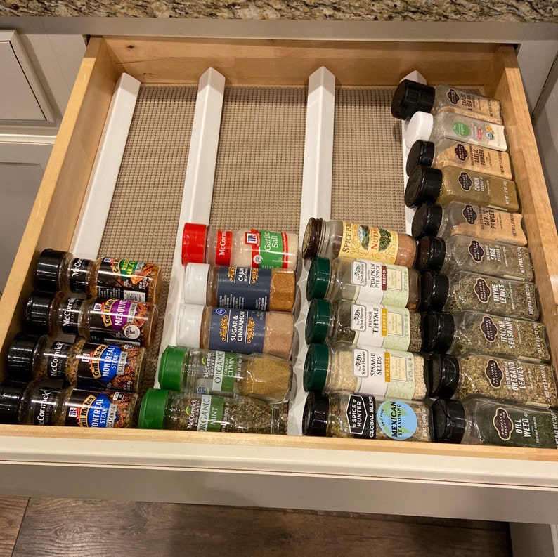 May include: A drawer organizer with white dividers holds a variety of spices in glass jars. The jars are labeled with different spice names, such as "Montreal Steak", "Sugar & Cinnamon", "Thyme", "Dill Weed", and "Italian Oregano".