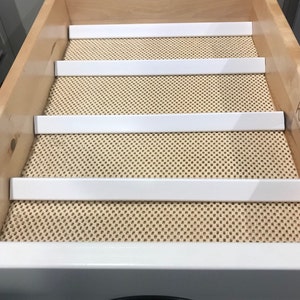 以下が含まれることがあります： キッチン用品を整理するために設計された、4つの仕切りが付いた白い引き出しライナー。ライナーは、ベージュのテクスチャ加工された引き出しマットの上に置かれています。