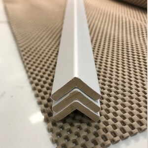 May include: Three white corner pieces stacked on top of each other. The corner pieces are made of a hard, smooth material and have a slightly rounded edge. The corner pieces are sitting on a brown, textured surface.