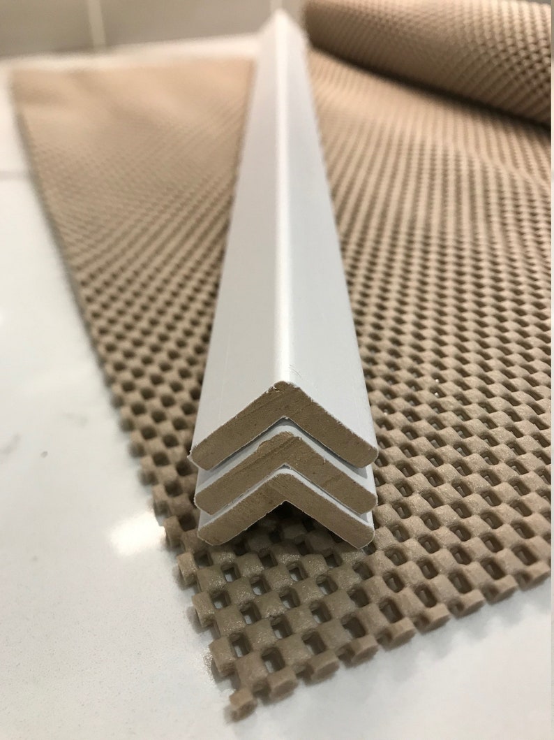 May include: Three white corner pieces stacked on top of each other. The corner pieces are made of a hard, smooth material and have a slightly rounded edge. The corner pieces are sitting on a brown, textured surface.
