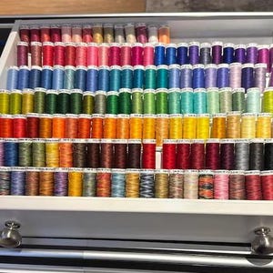 May include: A drawer filled with spools of colourful thread, organised by colour. The thread is arranged in rows, with each row containing a different colour. The colours include red, orange, yellow, green, blue, purple, and pink.