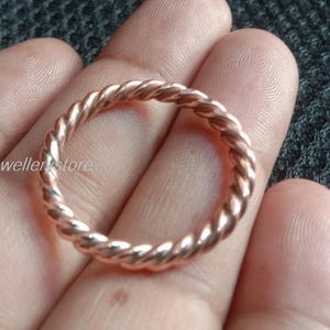 Könnte beinhalten: Ein roségoldfarbener Ring mit einem gedrehten Seildesign. Der Ring ist kreisförmig und liegt auf einer Hand, mit einem dunkelgrauen Hintergrund. Der Ring ist ein Schmuckstück.