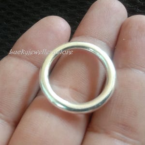 Könnte beinhalten: Ein silberner Ring mit glattem, abgerundetem Design. Der Ring wird in einer Hand gehalten, und die Oberfläche des Rings reflektiert das Licht. Der Ring ist ein schlichtes, elegantes Schmuckstück.