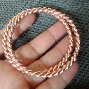 Op de afbeelding: Twee koperkleurige, gedraaide armbanden in touwstijl worden in een hand gehouden. De armbanden zijn gemaakt van een glanzend metaal en hebben een gevlochten uiterlijk. De achtergrond is een grijze stof.