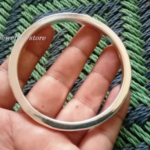 Könnte beinhalten: Ein silberfarbenes, kreisförmiges Armreif-Armband. Das Armband wird in einer Hand gehalten, mit dem Text "Lucky jewel store" sichtbar. Der Hintergrund ist ein gewebtes Muster aus Grün, Blau und Schwarz.