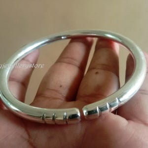 Könnte beinhalten: Ein silberfarbenes, offenes Armreif-Armband. Das Armband hat ein glattes, abgerundetes Design mit einer Reihe rechteckiger Vertiefungen in der Nähe der Öffnung. Das Armband wird in einer Hand gehalten.