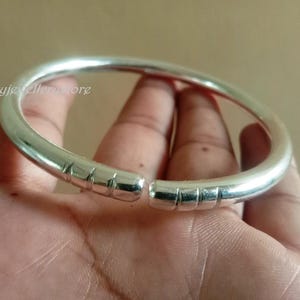 Könnte beinhalten: Ein silberfarbenes, offenes Armreif-Armband. Das Armband hat ein glattes, abgerundetes Design mit einer Reihe rechteckiger Vertiefungen in der Nähe der offenen Enden. Das Armband wird in einer Hand gehalten.