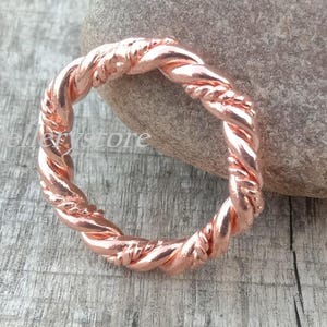 Könnte beinhalten: Ein kupferfarbener Ring mit einem gedrehten Seildesign. Der Ring ist kreisförmig und liegt auf einer verwitterten Holzoberfläche neben einem glatten, grauen Stein. Die Textur und Farbe des Rings werden hervorgehoben.
