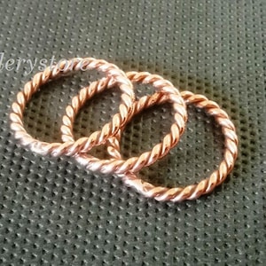 Könnte beinhalten: Drei kupferfarbene Ringe im gedrehten Seil-Stil werden auf einer strukturierten, dunkelgrünen Oberfläche gezeigt. Die Ringe sind in einer lockeren Anordnung angeordnet, mit dem Text "Luckyjewelleryst" oben links.