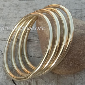 May include: A set of four gold-colored bangles resting on a brown stone. The bangles are circular and have a smooth, polished finish. The background is a weathered wooden surface.