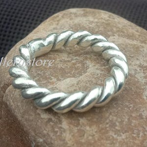May include: A silver ring featuring a twisted rope design. The ring is circular and rests on a light brown stone. The text "Luckyjewellerstore" is visible in the image.