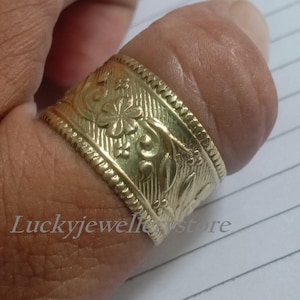 Könnte beinhalten: Ein goldfarbener Ring mit einem aufwendigen Blumen- und Schriftrollenmuster. Der Ring ist breit und umschließt den Finger. Der Ring befindet sich an einer Hand, und der Text "luckyjewellerstore" ist sichtbar.