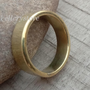 Könnte beinhalten: Ein goldfarbener Ring mit flachem Band. Der Ring liegt auf einem hellen Stein und einer Holzoberfläche. Der Ring ist ein schlichtes, klassisches Design.