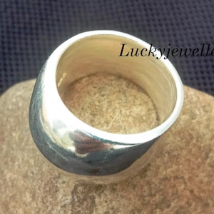 Könnte beinhalten: Ein breiter, silberfarbener Ring mit glatter, polierter Oberfläche. Der Ring wird auf einem hellbraunen Stein vor einem dunkelblauen, strukturierten Hintergrund gezeigt. Der Text "Luckyjewellerystore" ist auf dem Ring zu sehen.