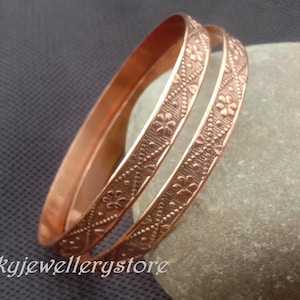 Könnte beinhalten: Zwei kupferfarbene Armreifen mit floralen und geometrischen Mustern. Die Armbänder liegen auf einem grauen Stein, mit dem Text "luckyjewellerystore" unten links.