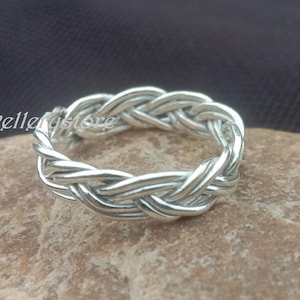Könnte beinhalten: Ein silberfarbener Ring mit geflochtenem Design. Der Ring besteht aus miteinander verflochtenen Strängen, wodurch ein strukturiertes, gewebtes Aussehen entsteht. Der Ring wird auf einer hellbraunen Steinoberfläche mit dunklem Hintergrund gezeigt.