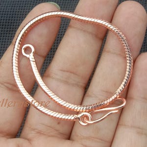 Könnte beinhalten: Ein roségoldenes Schlangenkettenarmband mit Hakenverschluss. Das Armband ist kreisförmig und flexibel, mit einer strukturierten Oberfläche. Der Verschluss ist ein einfaches Haken- und Ösendesign. Das Armband wird vor einem dunkelgrauen Hintergrund gezeigt.