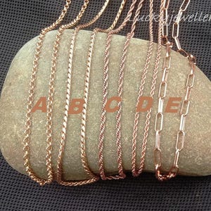 Könnte beinhalten: Eine Auswahl an roségoldenen Halsketten mit verschiedenen Kettendesigns, präsentiert auf einem hellbraunen Stein. Die Kettenarten umfassen Seil-, Curb- und Paperclip-Designs. Der Hintergrund ist ein dunkelgraues, strukturiertes Gewebe.