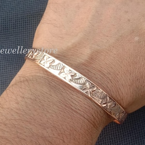 Op de afbeelding: Een koperkleurige manchetarmband met een gegraveerd bloemmotief. De armband wordt om een pols gedragen en het ontwerp bevat gestileerde bloemen en bladeren. De armband is ongeveer 2 cm breed.