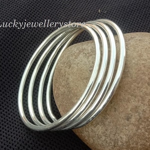 Könnte beinhalten: Fünf silberne Armreifen liegen auf einem hellbraunen Stein vor einem schwarzen, strukturierten Hintergrund. Die glatten, glänzenden Armreifen reflektieren das Licht. Der Text "Luckyjewellerystore" ist sichtbar.