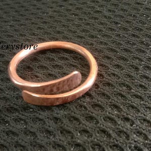 Könnte beinhalten: Ein kupferfarbener Wickelring mit gehammerter Textur. Der Ring hat ein offenes Design, wobei sich ein Ende überlappt. Der Ring wird auf einer dunklen, strukturierten Oberfläche präsentiert. Der Text "luckyjewellerystore" ist sichtbar.