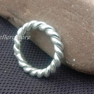 Pode incluir: Um anel prateado com um design de corda torcida. O anel está sobre uma pedra castanha, sobre um fundo texturizado cinza escuro. O texto "luckyjewellerystore" é visível.