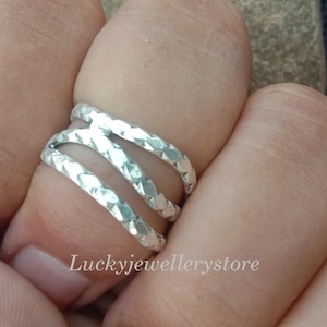 Könnte beinhalten: Ein silberner Ring mit einem strukturierten, überlappenden Design. Der Ring hat drei Bänder, die sich kreuzen und einen einzigartigen und modernen Look erzeugen. Der Ring wird an einem Finger getragen und zeigt seine komplizierten Details.